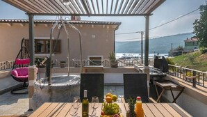 Uma mesa de madeira posta com copos de vinho, um prato de frutas e uma garrafa de suco, sob uma pérgola com vista para o mar.