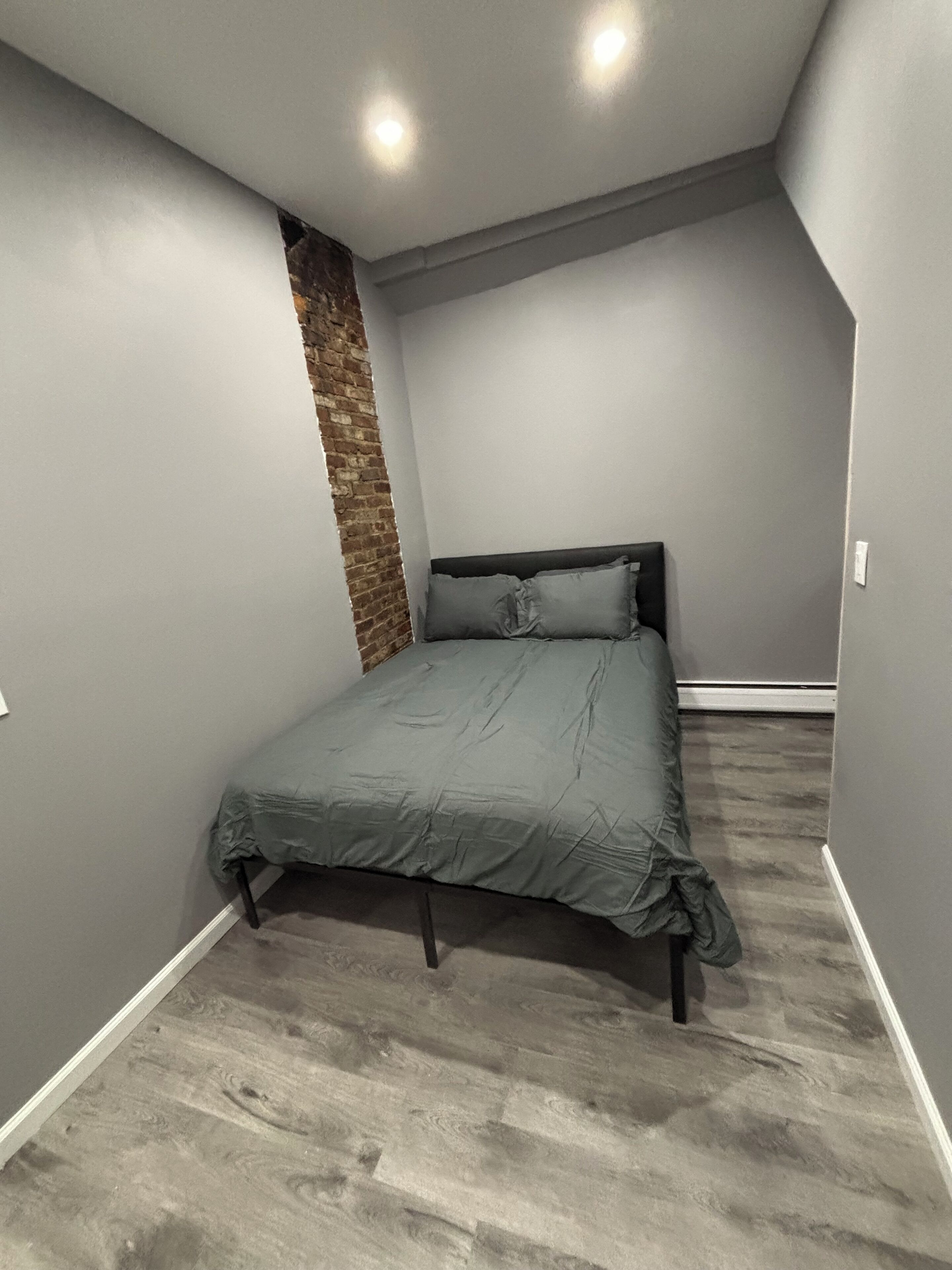 Ein Schlafzimmer mit Bett, grauen Wänden, einer Wand aus Backsteinen und einer schrägen Decke.