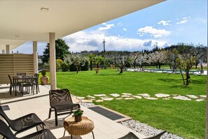 Un porxo cobert amb mobles d'exterior, gespa i vista d'un parc amb arbres i un edifici al lluny.