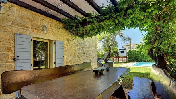 An outdoor dining area with a stone wall, wooden benches, and a view of a swimming pool. - Villa Sherpa (74136-K1) - Porec (Porec)
