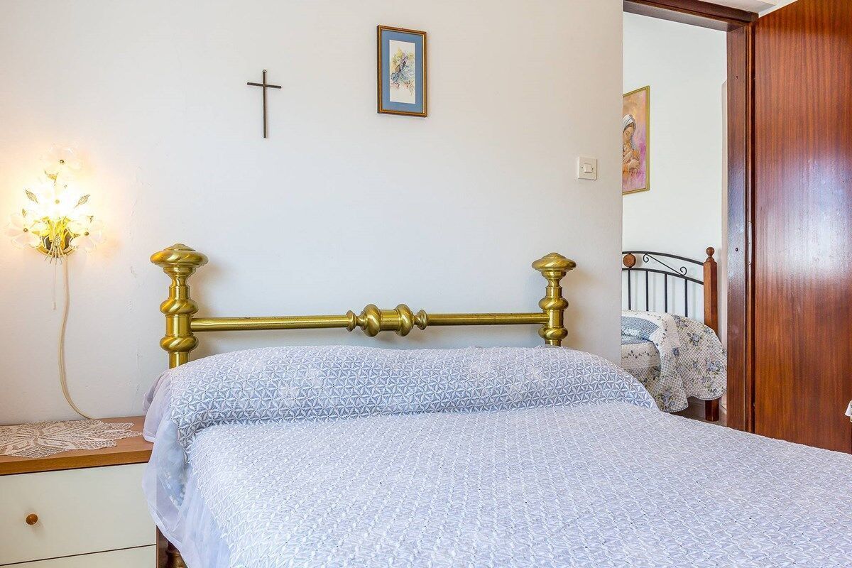 Ein Schlafzimmer mit Bett, Nachttisch, Lampe, einem Kruzifix an der Wand und einer Tür.