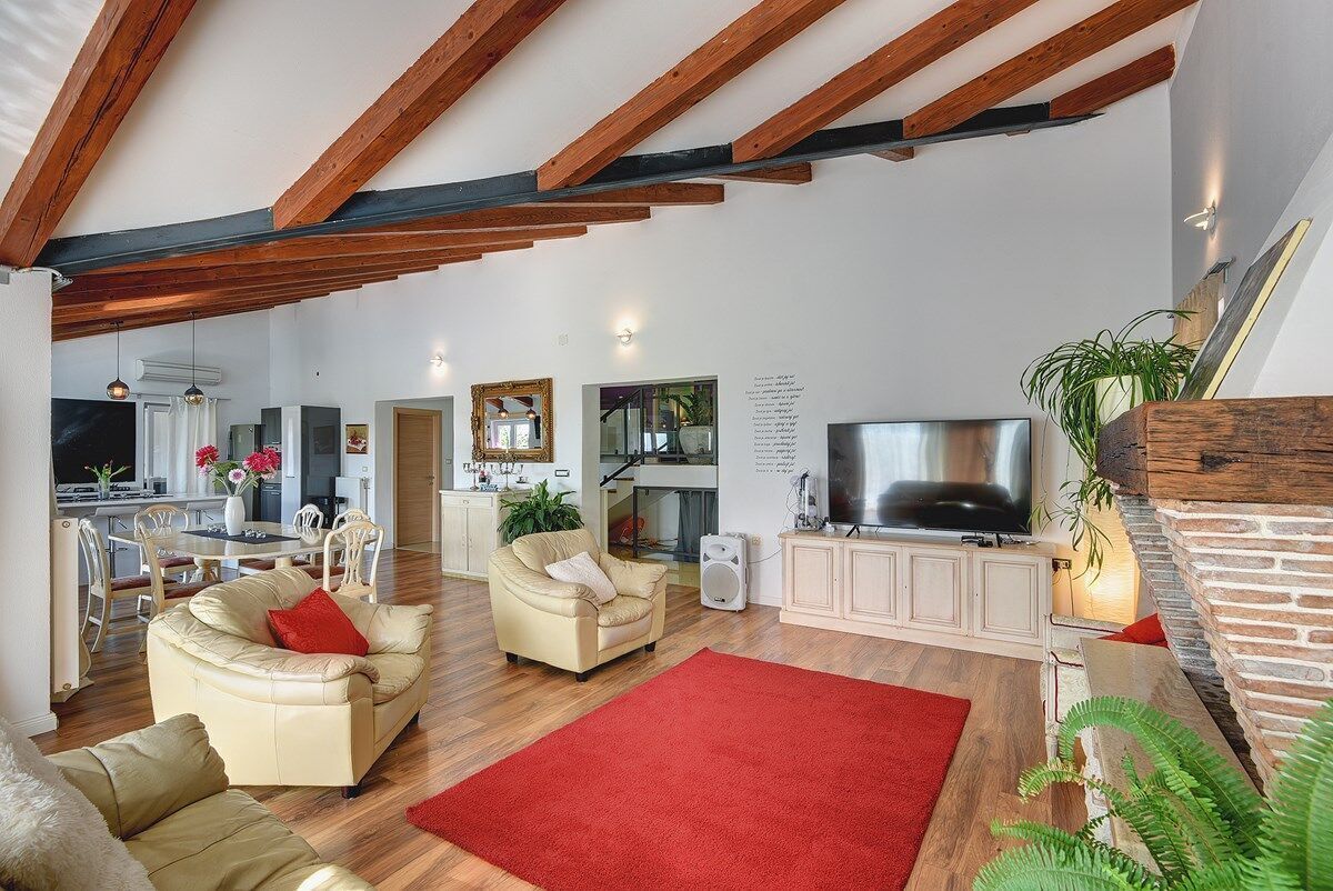 A spacious living room with a red rug, wooden beams, a TV, and a dining area in the background.