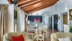 Uma sala de estar ampla com teto alto e vigas de madeira aparentes, uma área de jantar com mesa e cadeiras, e uma cozinha ao fundo.