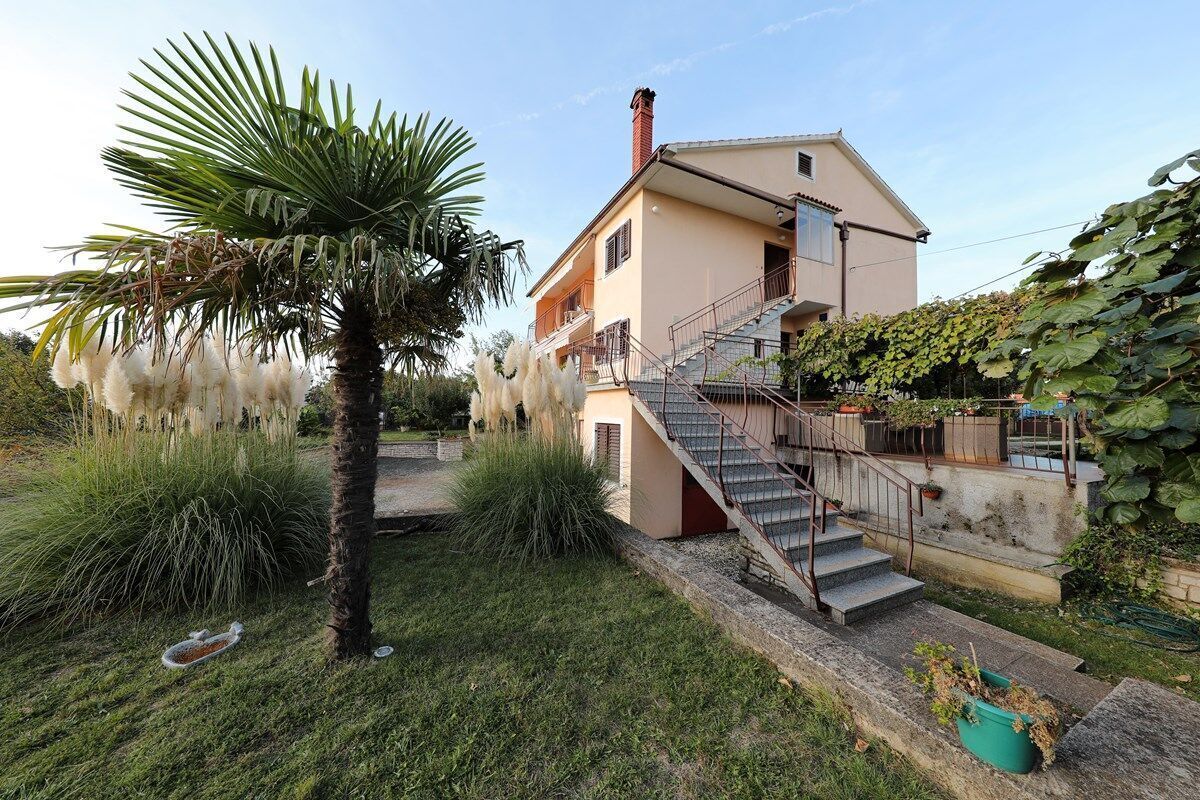 Kaksikerroksinen talo, jossa on portaikko, joka johtaa yläkertaan. Talon ympärillä on vehreää kasvillisuutta ja palmu.