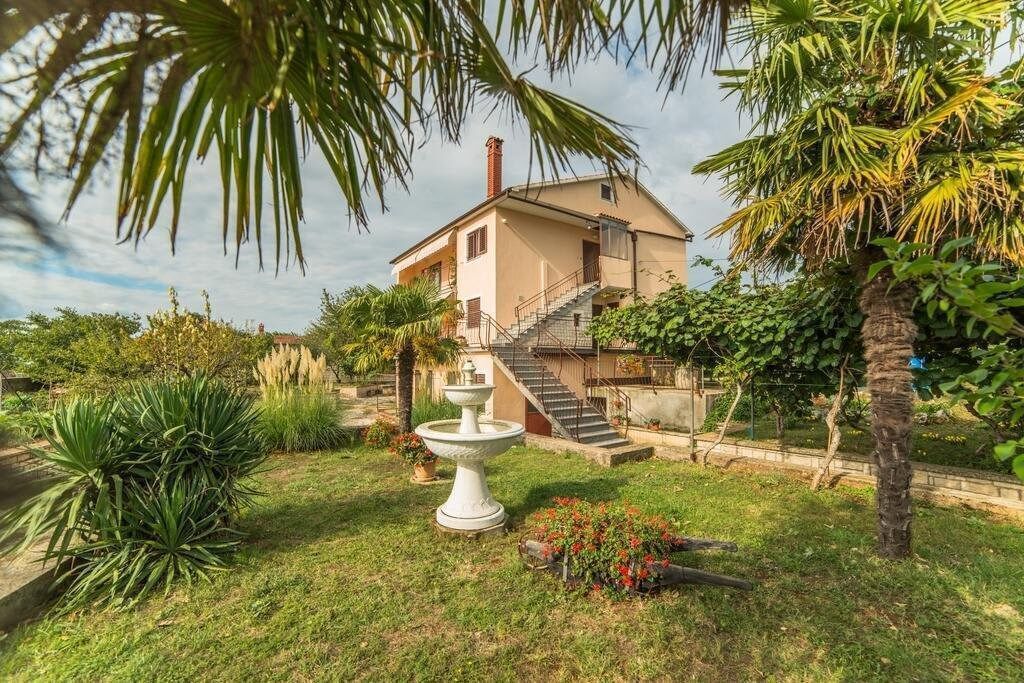 A two-story house with a staircase leading to the upper floor, surrounded by lush greenery and a white fountain.