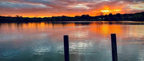 En træbro strækker sig ud i roligt vand, med en solnedgang i baggrunden.