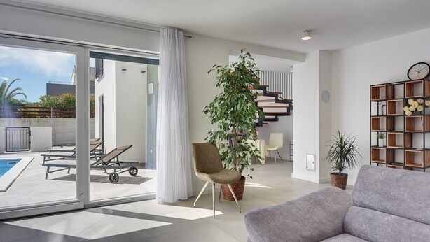 Ein modernes Wohnzimmer mit einem grauen Sofa, einer großen Kübelpflanze, einem Holzregal und einer Treppe, die zu einer weiteren Ebene führt.