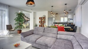 Ein modernes Wohnzimmer mit einer grauen Eckcouch, einem weißen Couchtisch und einer Topfpflanze. Eine Treppe führt zu einer oberen Ebene, es gibt eine Küchenzeile mit einem Esstisch und einen Kronleuchter.