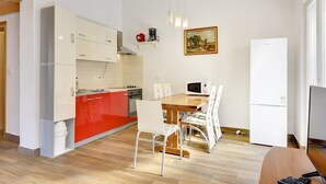 Ett kompakt kök med vita och röda skåp, ett matbord i trä med vita stolar, en mikrovågsugn och ett kylskåp.