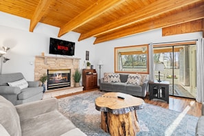 A cozy living room with a stone fireplace, a flat-screen TV, and a large window with a view of trees. - Carebear Cabin: Cowboy Game Room & Lake Access (Blakeslee)