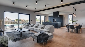A spacious living area with a large sofa, a coffee table, and a dining table with chairs. There is a kitchen area with dark cabinets and a hanging light fixture. - Villa Aquarius (74198-K1) - Pula (Pula)