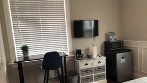 A bedroom with a bed, a desk with a chair, a television, a refrigerator, and a window with blinds.