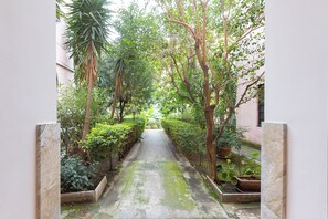 Un étroit sentier bordé d’une végétation luxuriante et de plantes en pot, qui mène à l’entrée d’un bâtiment.