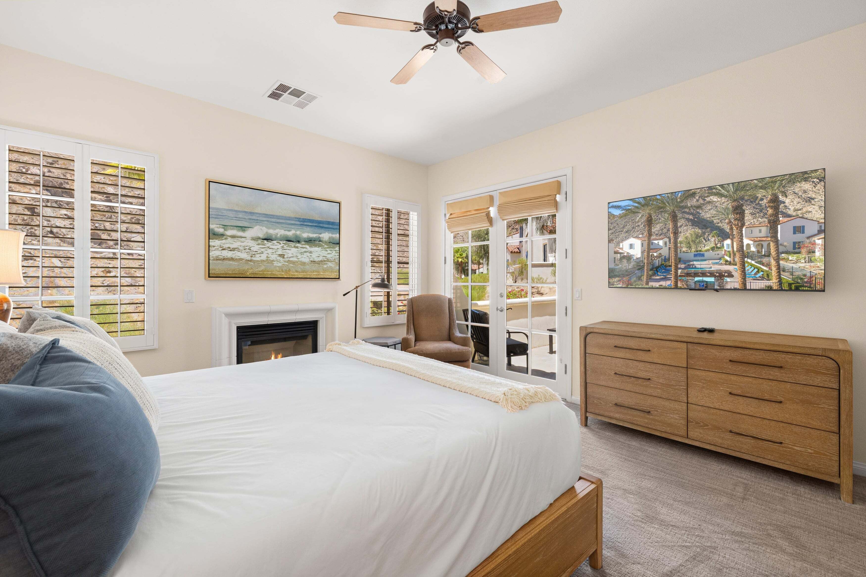 A bedroom with a bed, a fireplace, a ceiling fan, a dresser, and a large window with a view of a pool and palm trees.