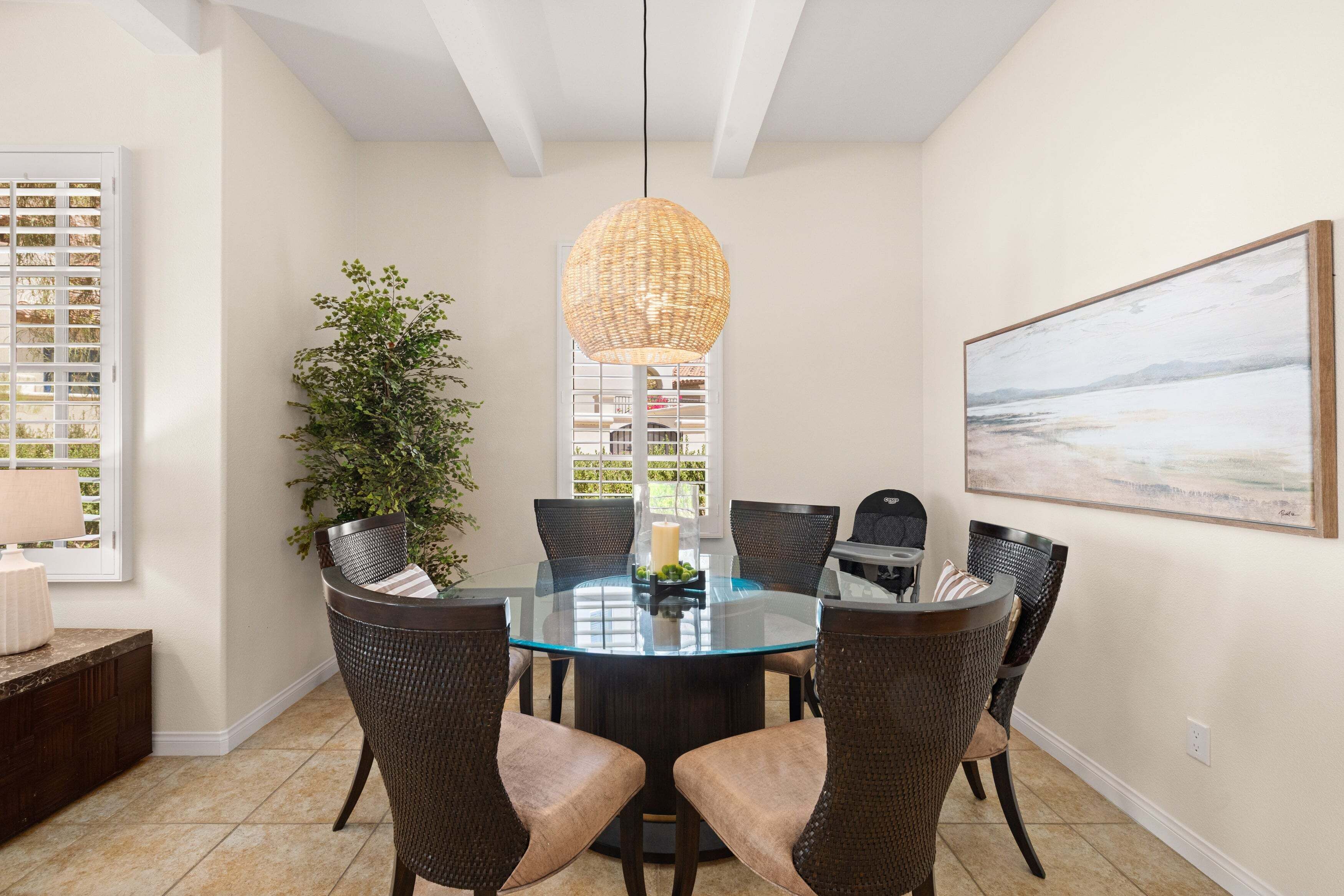 A dining area with a round glass table surrounded by wicker chairs, a hanging light fixture, and a painting on the wall.