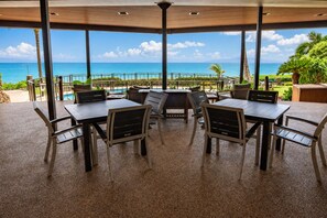 Ein überdachter Außenbereich zum Sitzen mit Tischen und Stühlen, mit Blick auf einen Pool und das Meer.