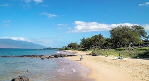 Ein Strand mit klarem, blauem Wasser, felsigem Ufer und einigen Spaziergängern.