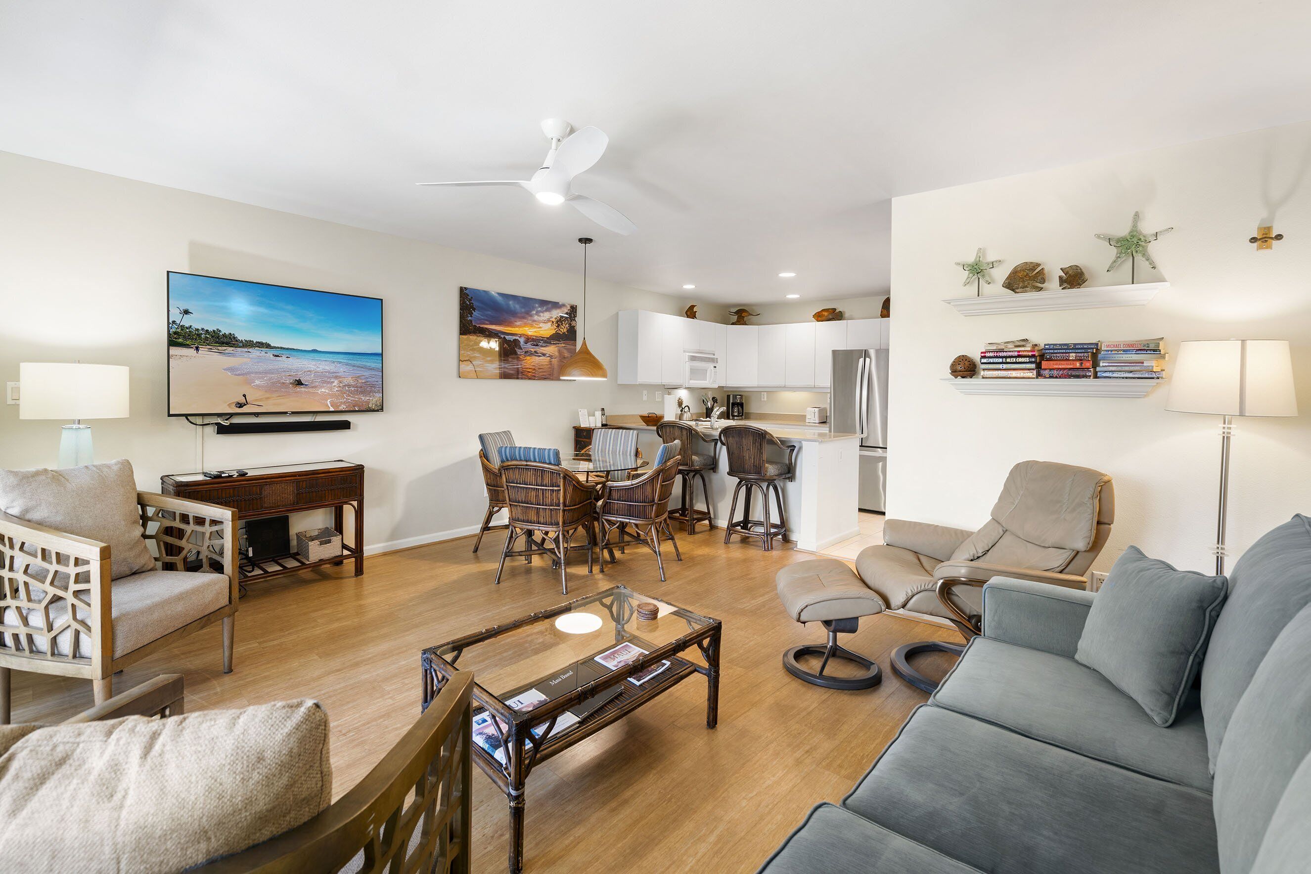 Un salon moderne comprenant un canapé, des fauteuils, une table basse et un coin repas avec une table et des chaises. Au fond, on aperçoit une cuisine avec des meubles blancs et un réfrigérateur.