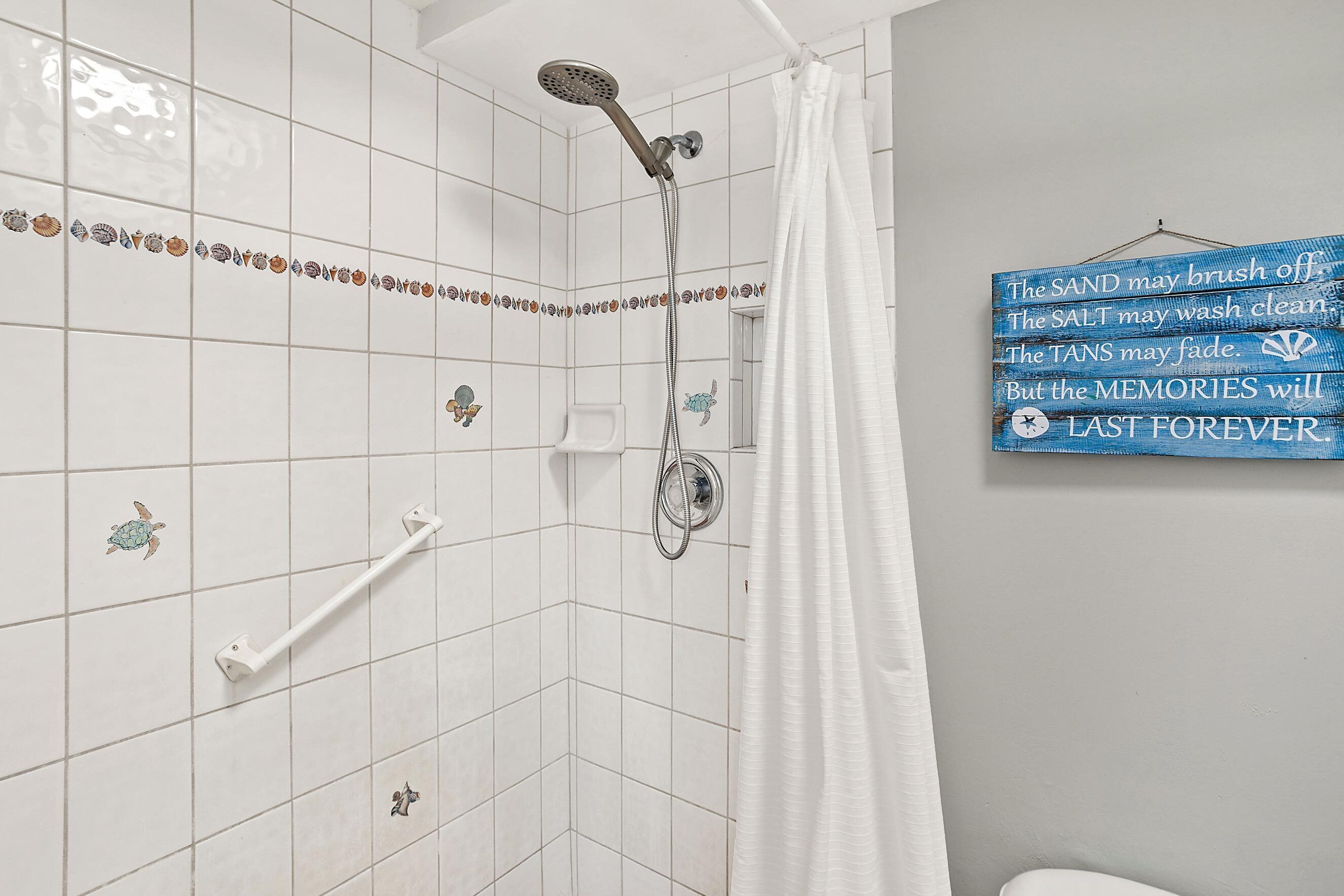 Een badkamer met witte tegels, een douche met handdouche, een witte handdoekhouder en een blauw bord met een citaat.