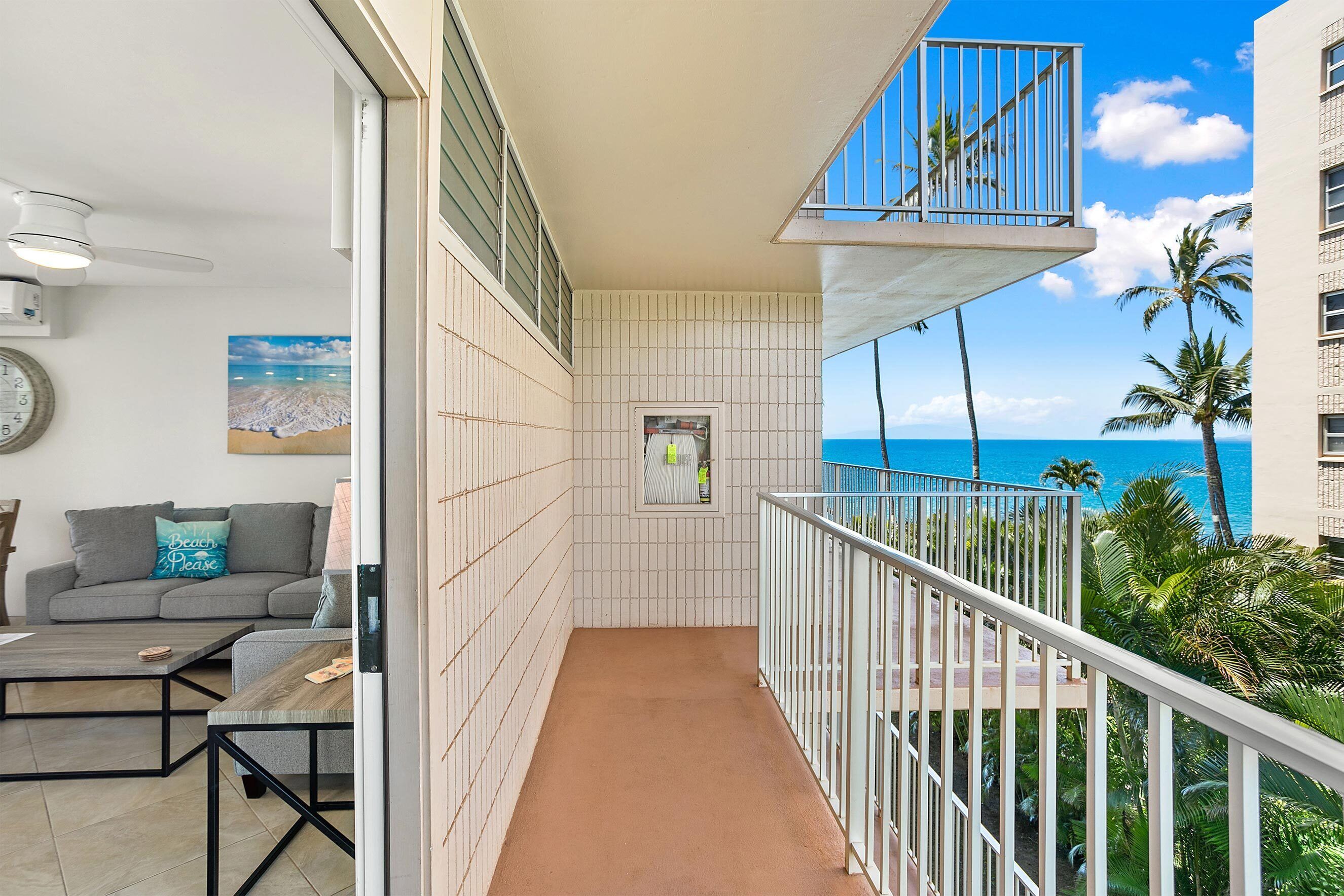 A balcony with a view of the ocean, a sofa with a 'Beach Please' pillow, and a small window with a picture.