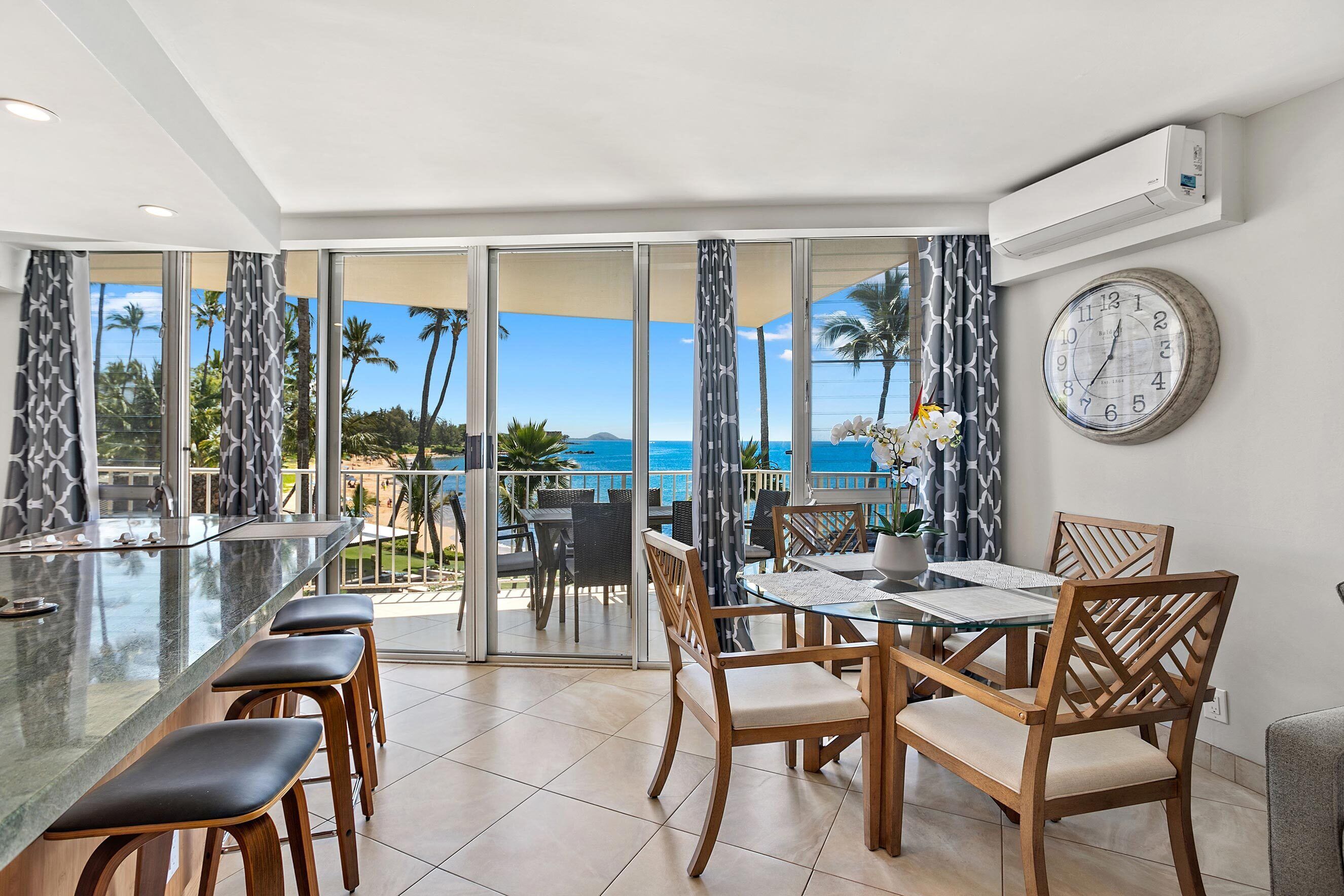 A dining area with a glass table and wooden chairs, a clock on the wall, and a view of the ocean through large windows.
