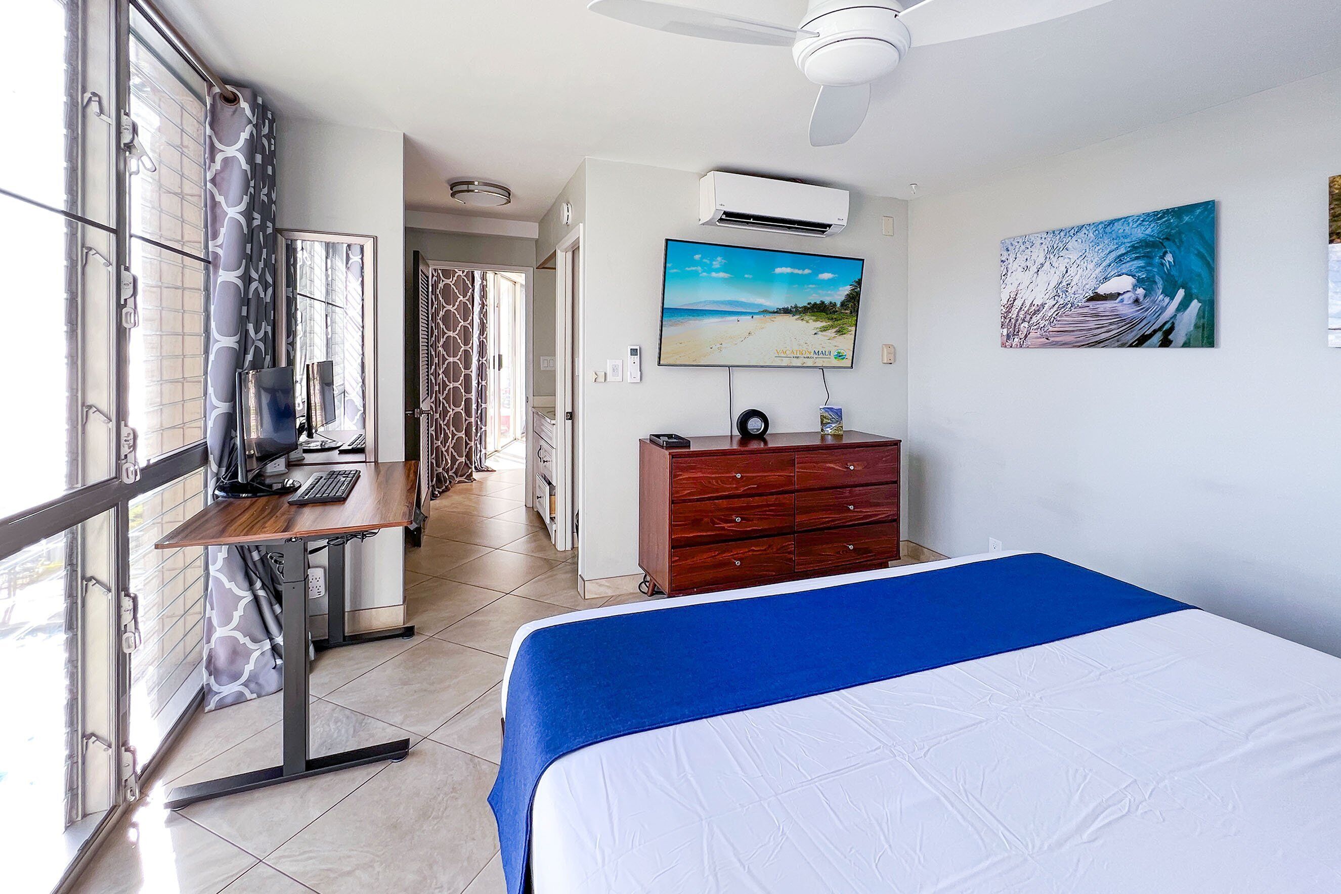 A hotel room with a bed, desk, and computer, a TV on the wall, and a window with curtains.