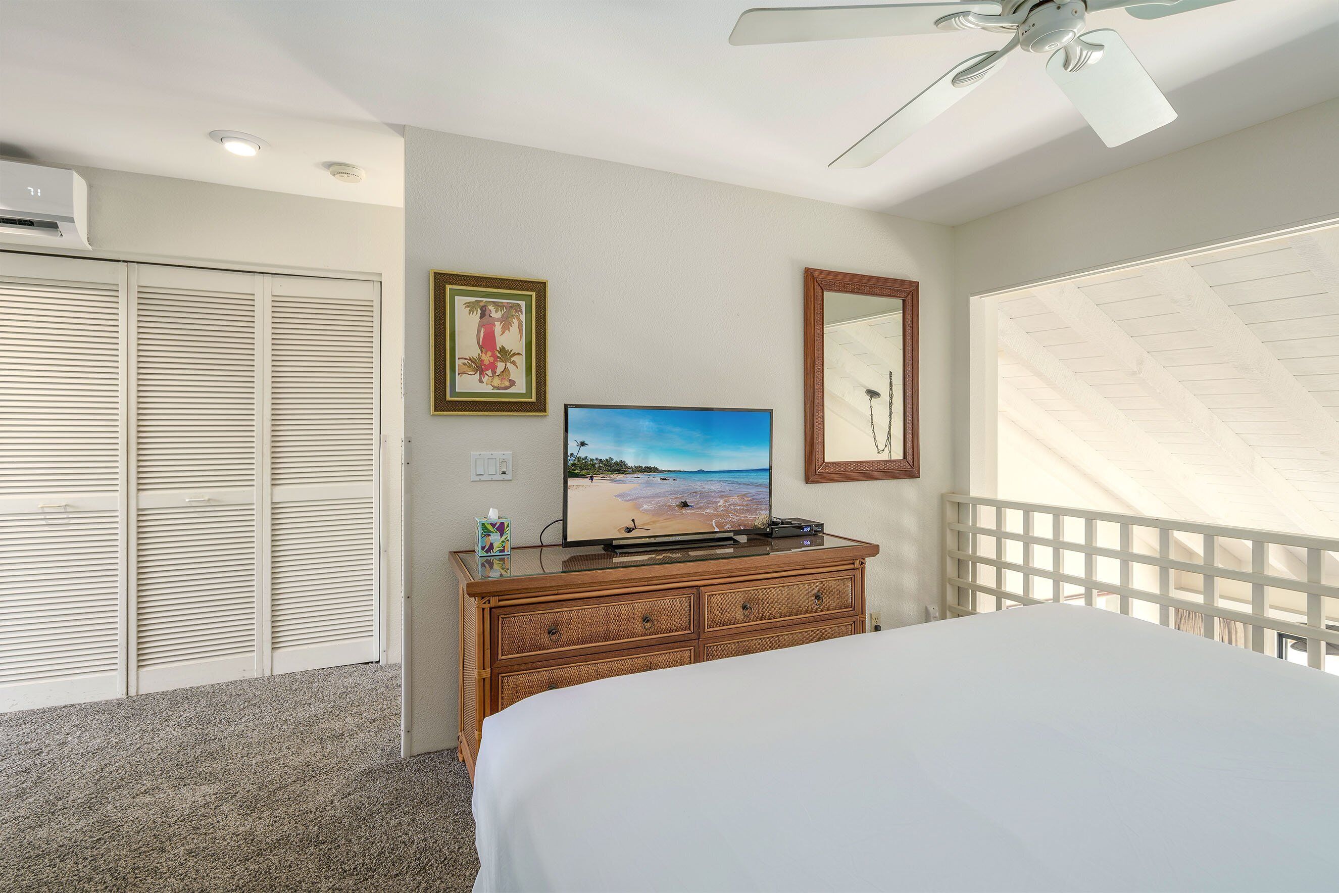 Un dormitorio con cama, televisor sobre una cómoda, ventilador de techo y vista a la playa a través de una ventana.