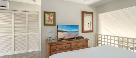 Un dormitorio con cama, televisor sobre una cómoda, ventilador de techo y vista a la playa a través de una ventana.