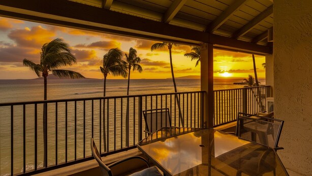 A balcony with a glass table and chairs, overlooking a sunset over the ocean with palm trees. - Lauloa 410 - Top Floor, Oceanfront Lanai, Molokini & Halaeakala Views, Split A/C (Wailuku)