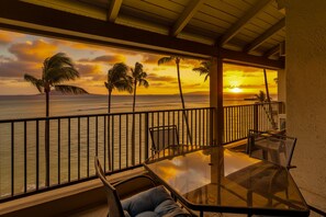 A balcony with a glass table and chairs, overlooking a sunset over the ocean with palm trees. - Lauloa 410 - Top Floor, Oceanfront Lanai, Molokini & Halaeakala Views, Split A/C (Wailuku)