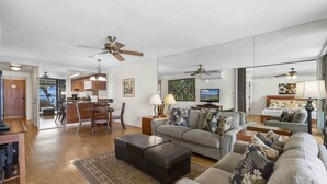 A spacious living area with a sofa, ottoman, and a dining table. There is a kitchen area with cabinets and a ceiling fan. - Lauloa 410 - Top Floor, Oceanfront Lanai, Molokini & Halaeakala Views, Split A/C (Wailuku)