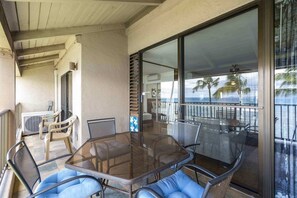 A covered outdoor area with a glass table and chairs, a view of the ocean, and a balcony with a railing. - Lauloa 410 - Top Floor, Oceanfront Lanai, Molokini & Halaeakala Views, Split A/C (Wailuku)