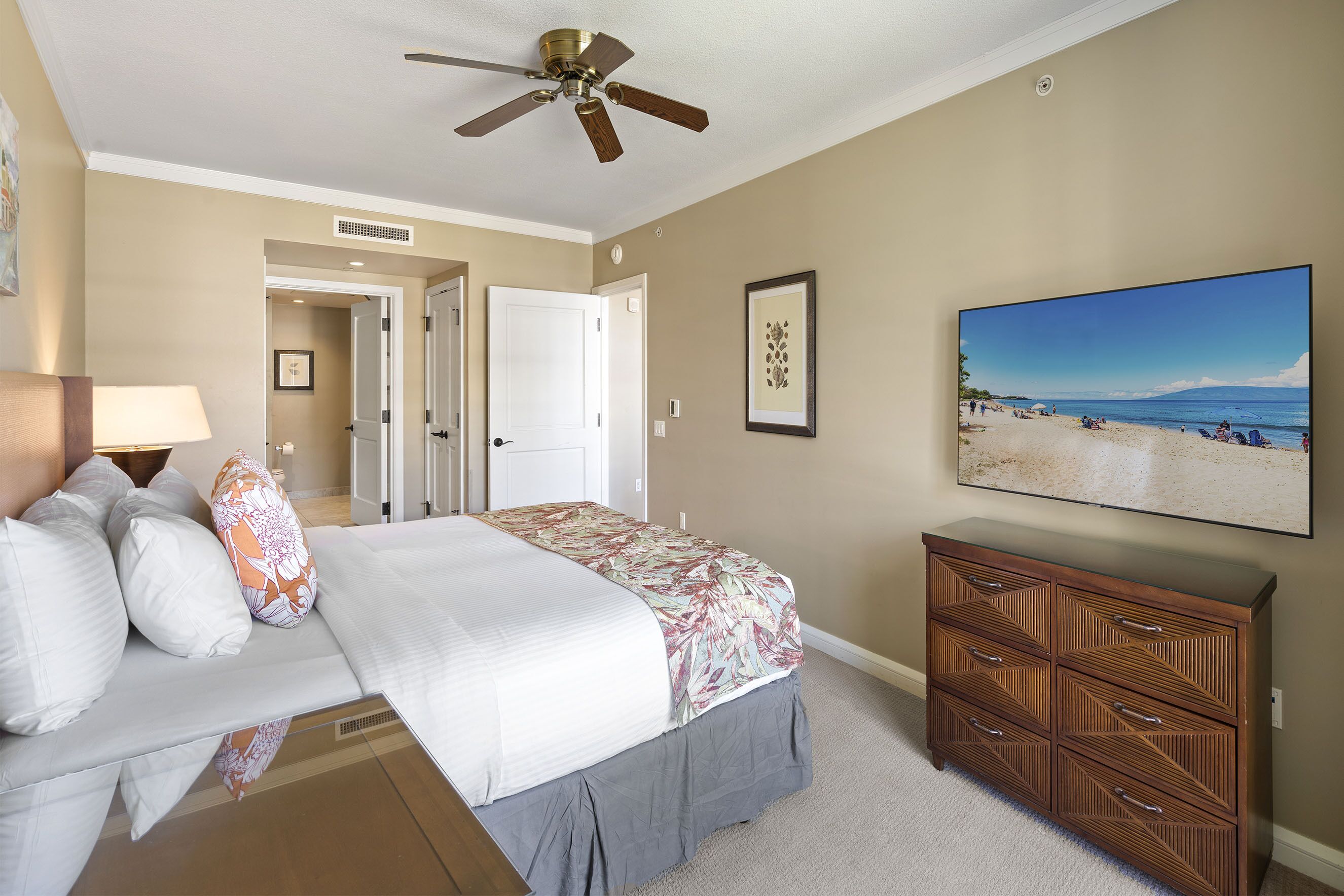 A bedroom with a bed, a ceiling fan, a dresser, a framed picture, and a painting of a beach.