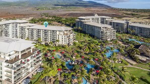 A resort with multiple high-rise buildings, a central pool area with palm trees, and a mountainous backdrop. - Honua Kai Resort Konea 505 Ocean & Sunset View 2BR at Kaanapali (Lahaina)