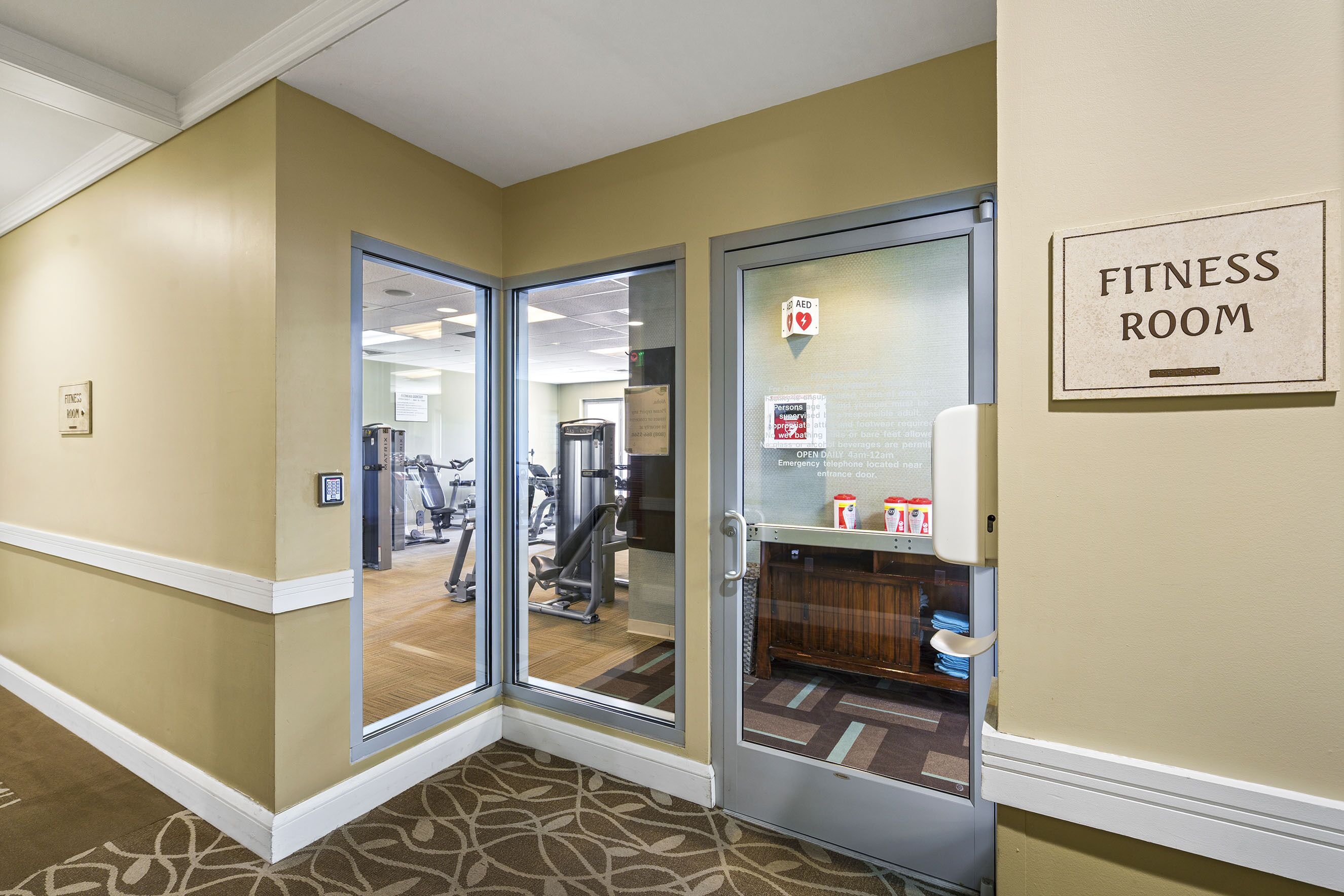 A fitness room with exercise equipment, a first aid sign, and a door leading to a carpeted hallway.