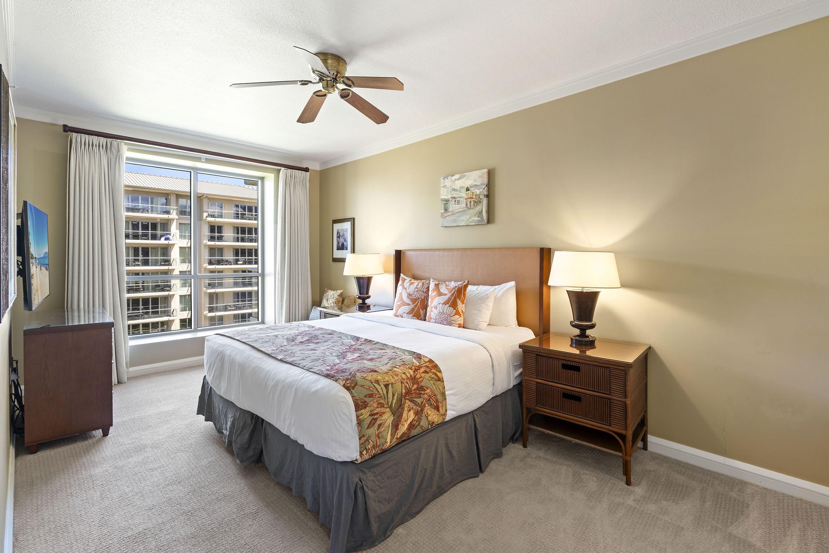 A hotel room with a large bed, a ceiling fan, a nightstand with a lamp, a dresser, and a window with curtains.