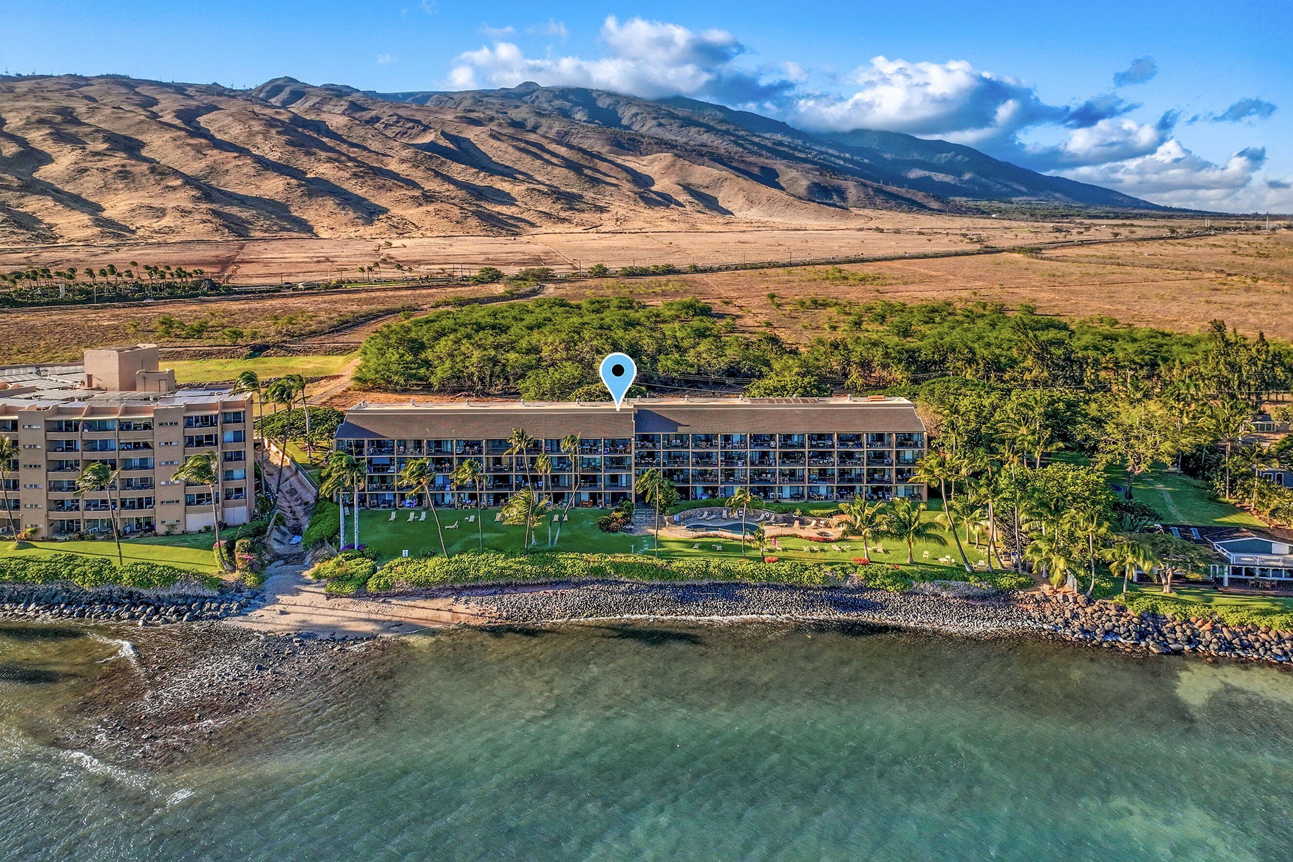 Une vue aérienne d’un complexe hôtelier avec une plage, une chaîne de montagnes en arrière-plan et un grand bâtiment sur lequel est apposée une épingle de localisation bleue.