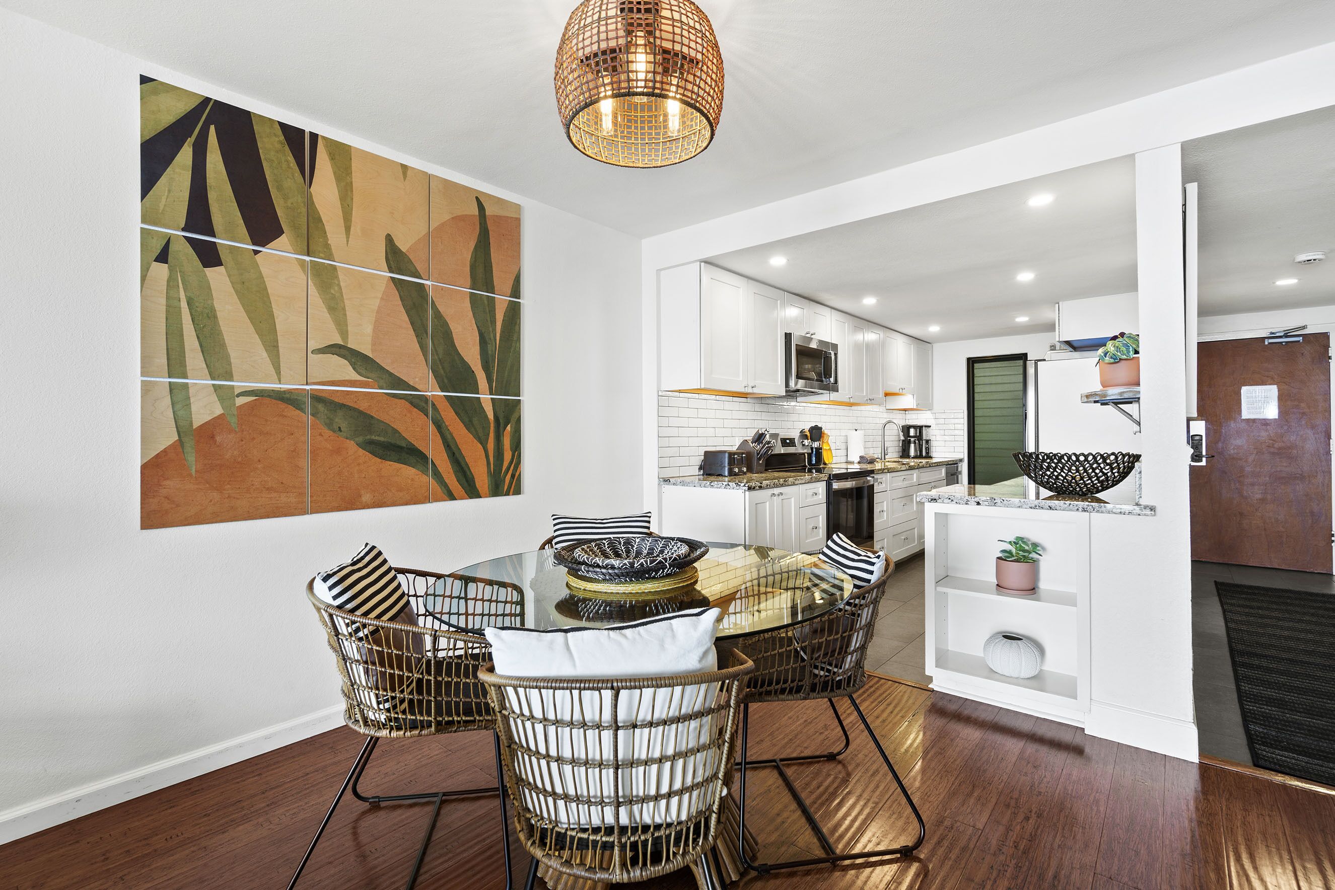 A modern kitchen with white cabinets, a glass dining table, wicker chairs, and a large abstract painting on the wall.
