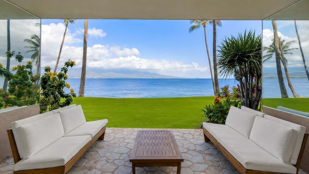 A patio with a white sofa, a wooden table, and a view of the ocean. - Oceanfront Lauloa 106 - Ground Floor, Modern Renovation, Split-ac, Walk to Beach (Wailuku)