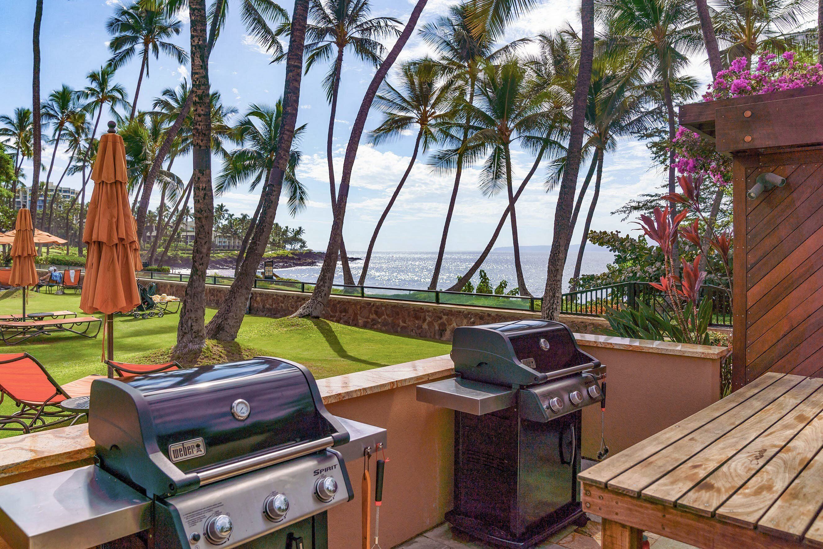 Eine Terrasse im Freien mit zwei Grills, einem Holztisch und Blick auf den Ozean mit Palmen.