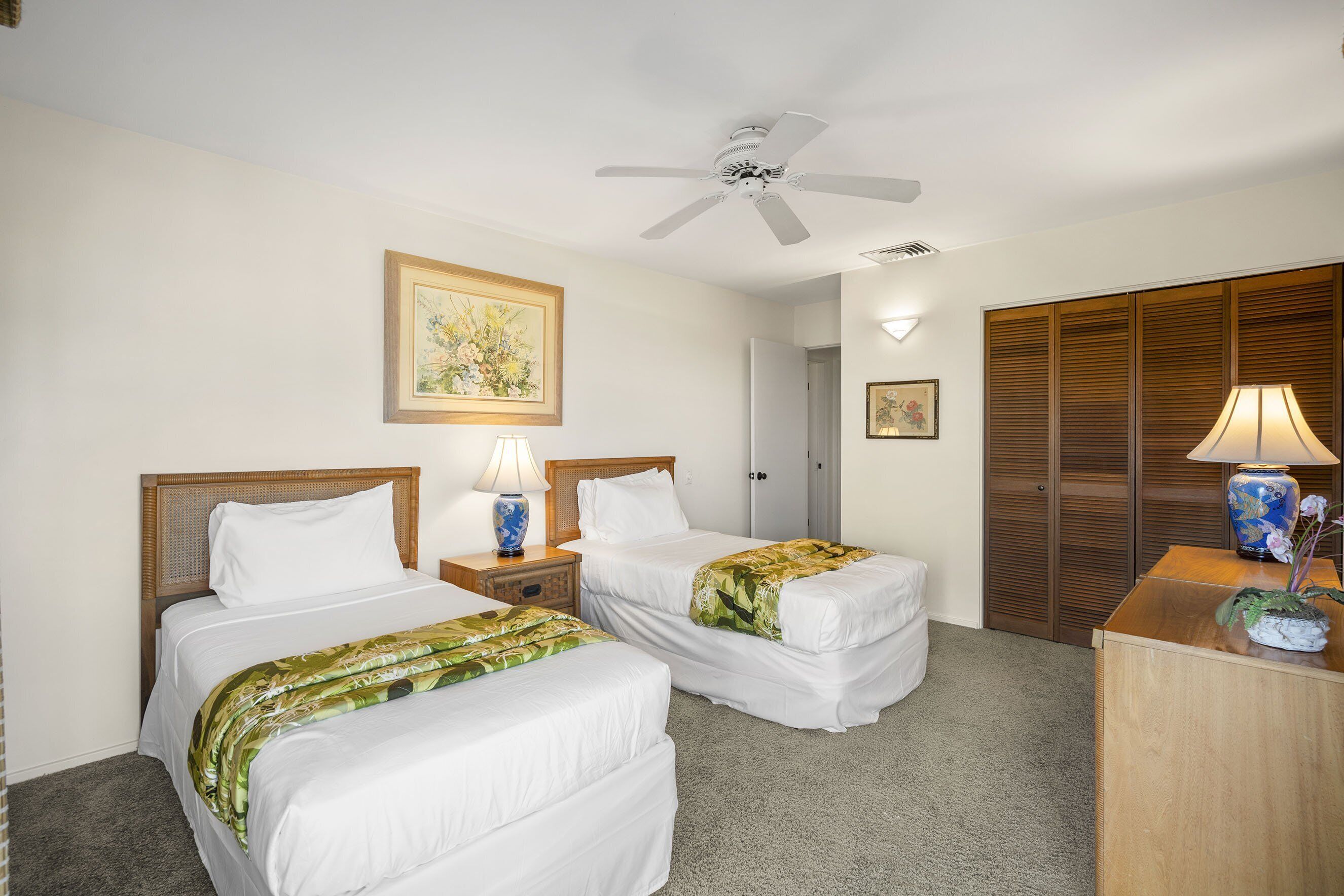 A hotel room with two beds, a ceiling fan, a wooden wardrobe, and a desk with a lamp.