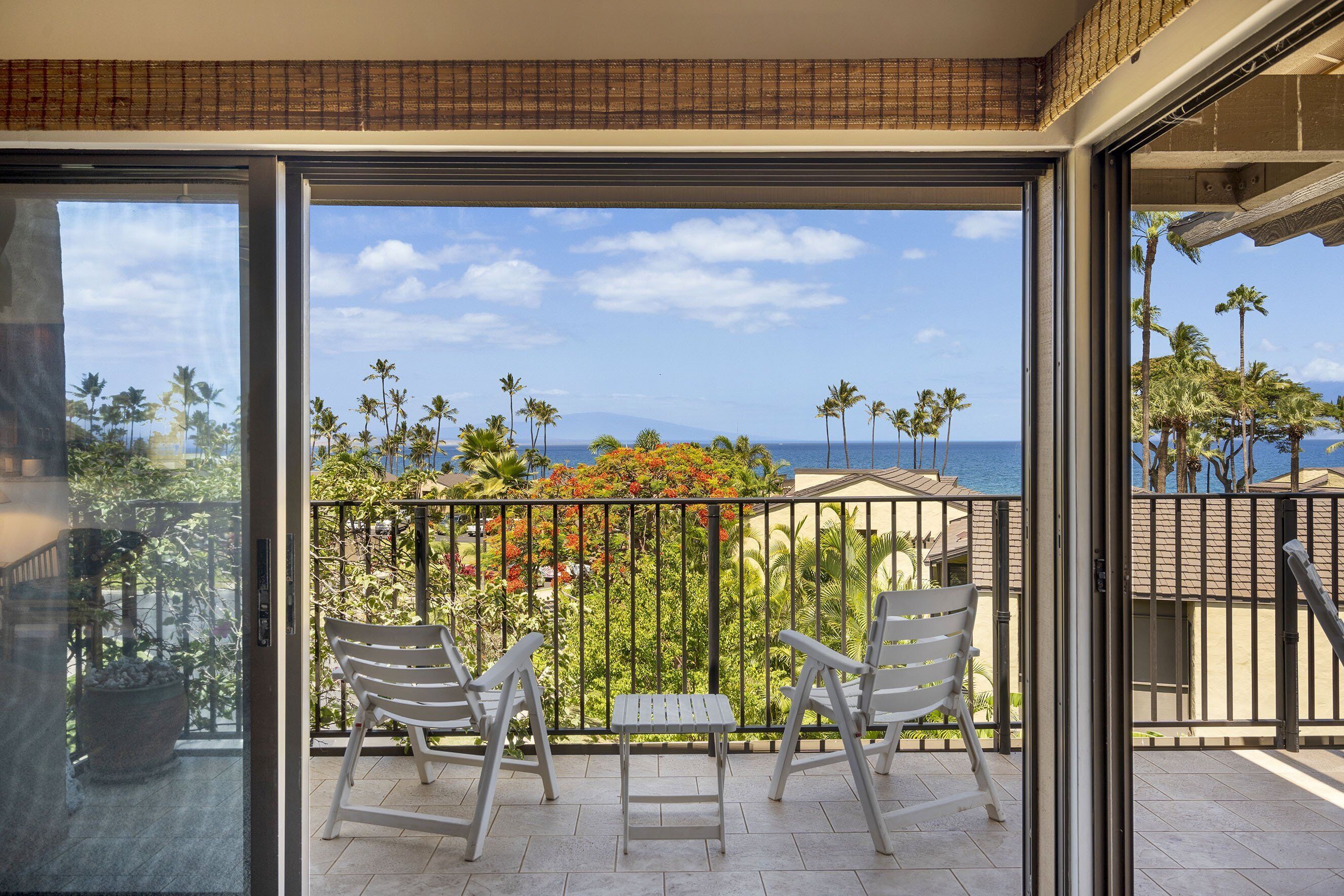 A balcony with two white chairs and a small table, overlooking a tropical landscape with palm trees and a clear sky.