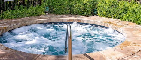 Un jacuzzi con un poste metálico en el centro, rodeado de baldosas de piedra, con un letrero y exuberante follaje al fondo.