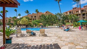 Zona de piscina en un resort con sillones de descanso y sombrillas, rodeada de palmeras y un edificio de varios pisos.