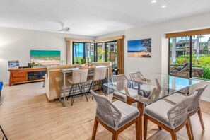 A modern dining area with a glass table and wicker chairs, a television, and artwork on the walls. - Luxurious Oceanfront Condo at Wailea Elua Village - Unit 1201, Central AC, Wi-fi (Kihei)