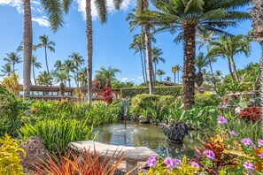 A tropical garden with a pond, palm trees, and a building with a modern design. - Luxurious Oceanfront Condo at Wailea Elua Village - Unit 1201, Central AC, Wi-fi (Kihei)