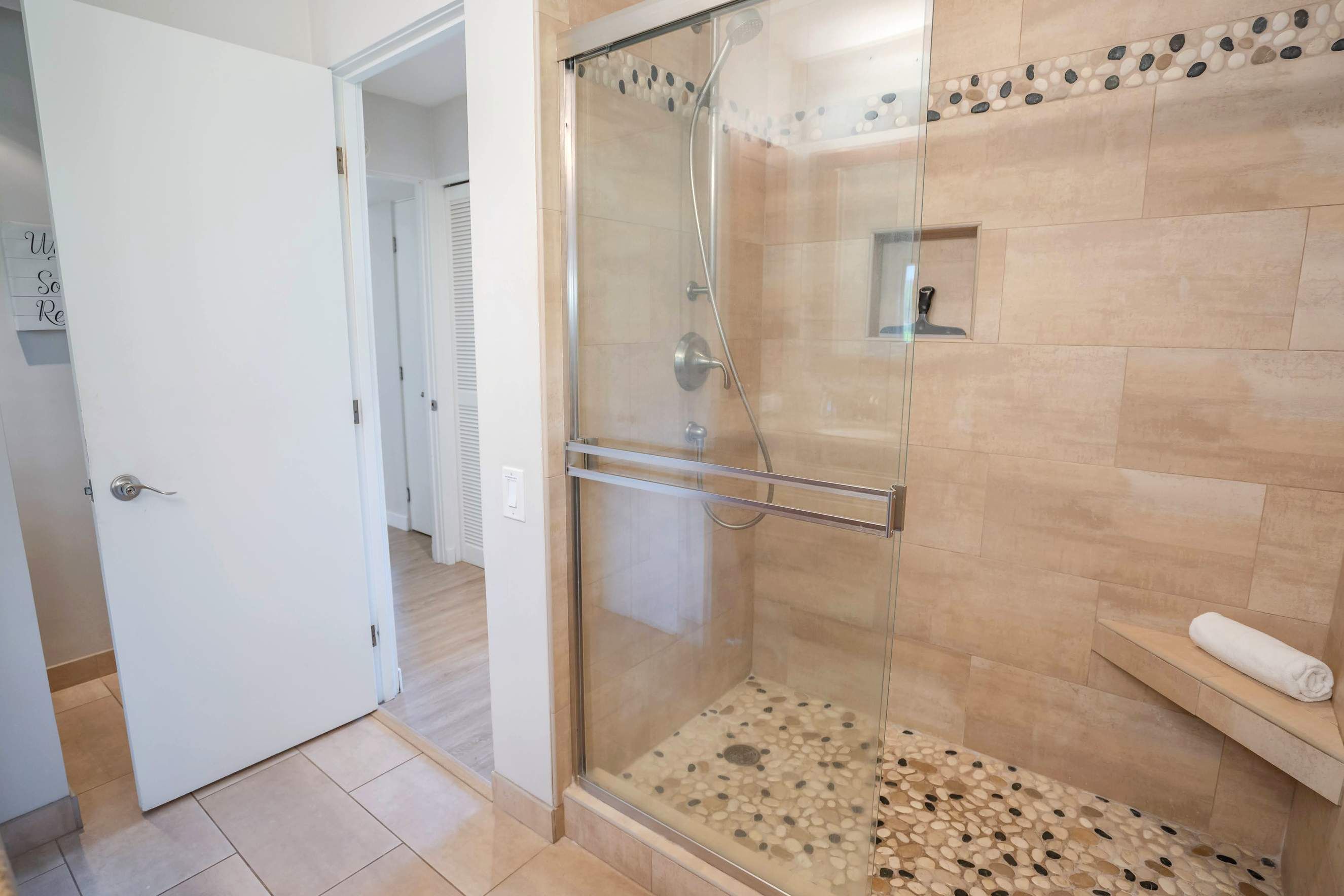 A glass-enclosed shower with a pebble-tiled floor, a towel rack, and a door leading to another room.