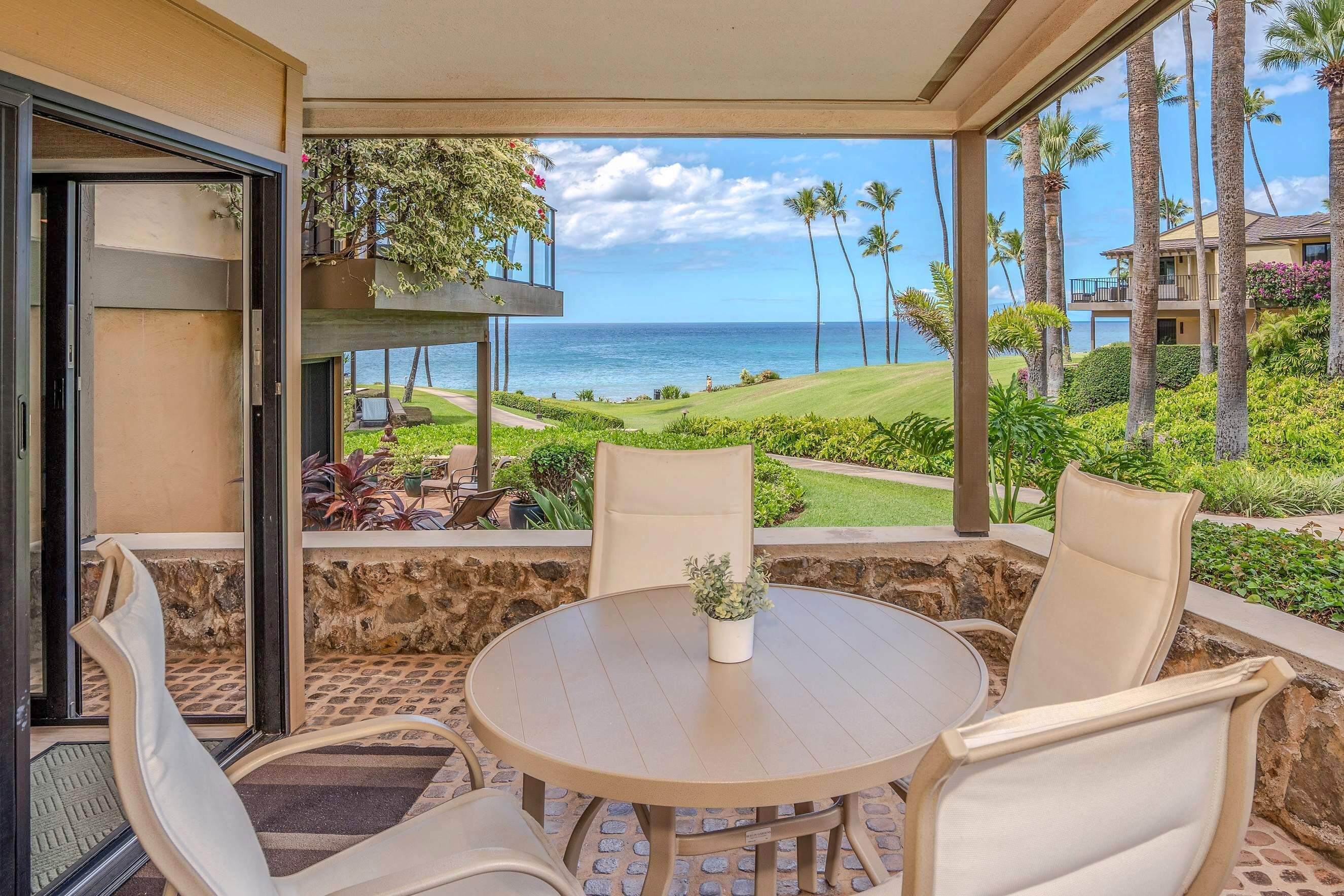 A covered outdoor seating area with a table and chairs, overlooking a view of the ocean and palm trees.