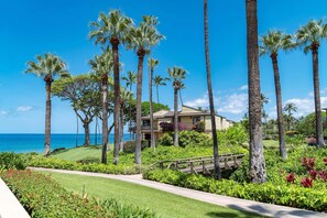 A beachfront property with a well-maintained lawn, palm trees, and a wooden bridge leading to a building with a balcony overlooking the ocean. - Luxurious Oceanfront Condo at Wailea Elua Village - Unit 1201, Central AC, Wi-fi (Kihei)
