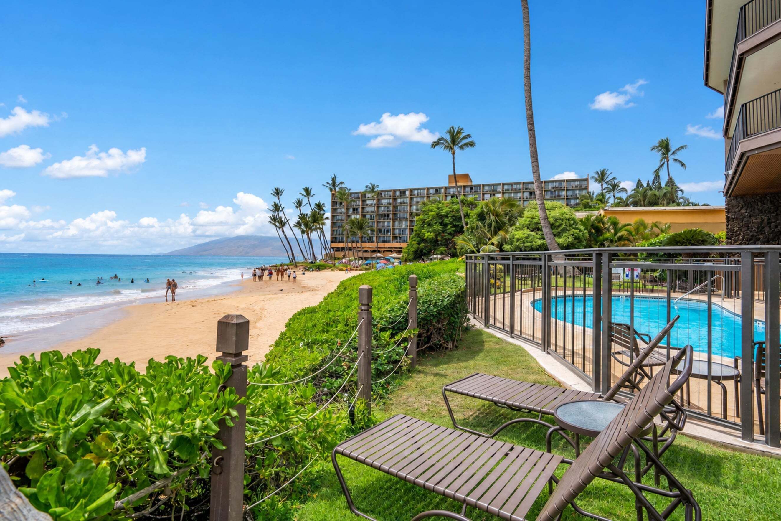 A poolside area with a view of the beach and ocean, surrounded by greenery and a building with multiple floors.